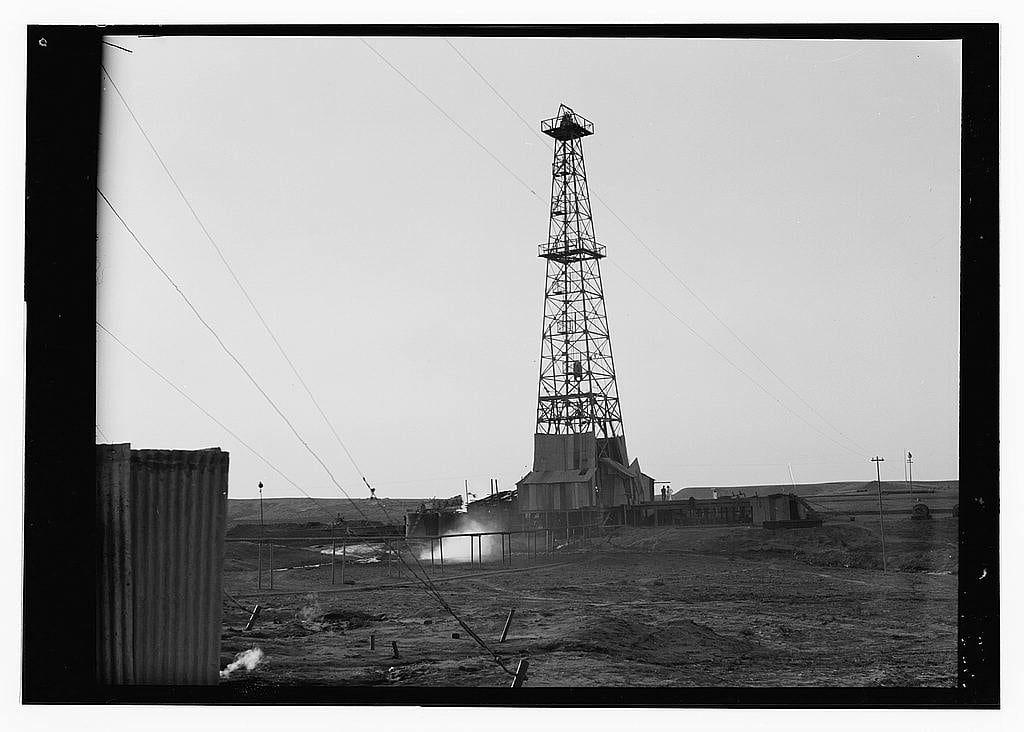 The image is a black and white photograph depicting an oil drilling tower in what appears to be an industrial or remote area. The tall, skeletal structure of the drill stands prominently against the overcast sky. Surrounding it are some simple buildings, possibly part of a temporary encampment for workers. There's minimal vegetation; barren land extends as far as one can see with hints of construction equipment and materials scattered around.

The photograph carries an air of desolation or hardship, common in early 20th-century industrial settings. The lighting suggests it could be either dawn or dusk due to the soft shadows cast by objects on the ground. There are no visible people; hence we cannot infer demographic details such as gender or race from this image.

Notable features include a corrugated metal structure and various other industrial equipment around, which indicate that these were operational oil fields during their time of operation likely in Iraq given historical context provided alongside the photo.