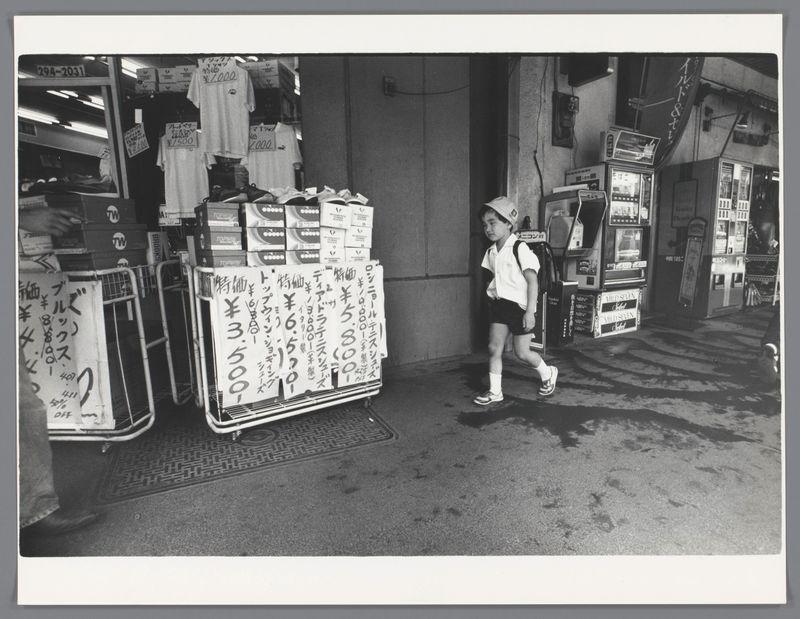 The image is a black and white photograph depicting an urban street scene, likely in Japan given the context provided. A young boy dressed in school uniform—white shirt with dark stripes on the sleeves, shorts, sneakers, and socks—is walking past a stall that appears to be selling various goods including t-shirts labeled "TW" and boxed items possibly for resale or distribution. The prices are written in Japanese yen, indicating currency usage specific to Japan.

The setting suggests an outdoor market area with assorted vending machines lined up on the right side of the frame displaying Japanese characters likely denoting drink offerings such as beer. In the foreground is a man partially visible from behind walking past the stall where the boy stands. The environment features a mix of concrete and tile flooring, common in urban pedestrian zones.

The photograph captures candid moments typically associated with street photography—a genre known for its unposed subjects and spontaneous capture of everyday life. This particular snapshot seems to be taken during daylight hours given the visible shadows on the ground. There is an airbrush effect applied over a large portion of the image which affects both foreground and background elements, suggesting that this photo might have been digitally altered or intentionally manipulated post-capture for artistic reasons.

The caption re [...]