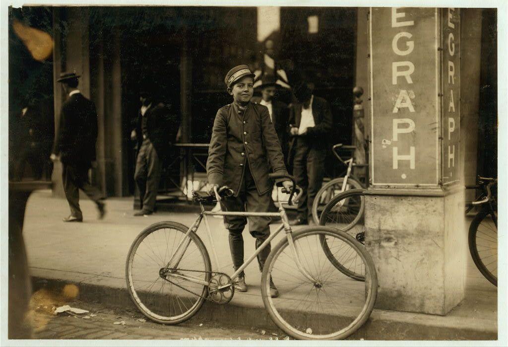 The photograph captures a young individual standing on the sidewalk with one foot near and the other resting against an old-fashioned bicycle. The person is dressed in what appears to be early 20th-century attire, consisting of a dark-colored coat adorned with buttons, possibly made from wool or another heavy fabric suitable for colder weather, paired with trousers that reach just above knee-length boots. They are wearing shoes and have their hands gripping the handlebars of the bicycle.

The street scene in the background features several individuals walking by, some appearing to be conversing with each other. The setting suggests an urban environment, possibly a downtown area given the presence of what appears to be a telegraph office or similar establishment labeled "EGRAPH" on a signpost behind them. Other bicycles are also visible further down the street.

The photograph is in black and white, which adds a historical context and indicates that it was likely taken during an earlier time period when color photography had not yet been widely adopted for everyday use. The overall atmosphere of the image suggests daily life as captured through documentary photography by Lewis Hine, who frequently photographed social reform issues such as child labor.

The specific reference to "Postal Telegraph messenger" in Birmingham, Alabama, hints at a connection between this photogra [...]