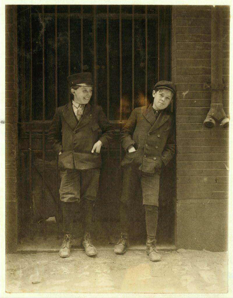 The image depicts two young boys dressed in early 20th-century work attire standing by a barred entrance. They appear to be workers, as suggested by their heavy coats and sturdy shoes, indicative of manual labor. The boy on the left wears a dark coat with buttons, trousers tucked into high boots, and is holding his hands loosely in his pockets while smiling at the camera. His companion stands slightly behind him, looking off to the side with a more contemplative expression; he also sports similar work attire including shoes appropriate for industrial settings.

The setting seems urban, possibly outside an industrial building given the barred door's metallic construction and robust brickwork visible on either side of it. There is no direct indication of their specific role at Inland Type Foundry or how long they have been working since "12th & Locust Sts." is not clearly legible in this context. However, based on historical records like those linked to Lewis Hine's work (a pioneering photojournalist known for documenting labor conditions), these boys likely represent the youth employed in such factories during that period.

This photograph exudes a sense of history and reflects early industrial working life where children were often involved in physically demanding jobs. The image captures their youthful innocence against an adult world backdrop, evoking thoughts on child  [...]