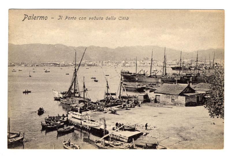 The image is a sepia-toned vintage postcard depicting the port of Palermo, Italy. It shows several traditional sailing ships and smaller boats docked at various piers within what appears to be an old harbor setting. The water is calm with few visible waves or ripples indicating peaceful weather conditions.

In the background, there are rolling hills or mountains that form a natural backdrop for the port area. Onshore structures include buildings and sheds typical of historical maritime facilities like warehouses or storage units possibly used during cargo handling operations in the past.

A handwritten inscription at the top reads "Palermo - Il Porto con veduta della Città," which translates to "Palermo - The Port with City View." This suggests that the postcard aims to showcase both the harbor and a view of Palermo's cityscape. In the bottom right corner, there is a signature or mark that appears faded but could be an indication of authorship or ownership.

The overall atmosphere conveyed by this image evokes a sense of history and maritime activity in Palermo from earlier times, potentially between late 19th to early 20th centuries when such sailing vessels were prevalent.