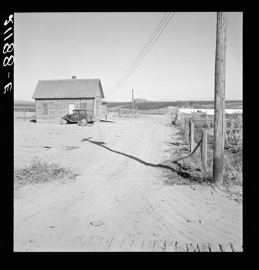 The image is a black and white photograph depicting an empty rural road leading towards a rustic wooden house with a weathered roof. To the left of the path, there's an old truck parked beside the building. The ground appears to be dry dirt with visible tire tracks indicating recent use. On either side of the road are barbed wire fences that demarcate property boundaries. A telephone pole stands prominently in the foreground on the right-hand side. In the distance beyond the house and fence, agricultural fields or farmland can be seen under a clear sky. The image carries historical connotations as it reflects a setting from rural America's past with its emphasis on simplicity and hard work through such details as an aged truck and wooden structures.