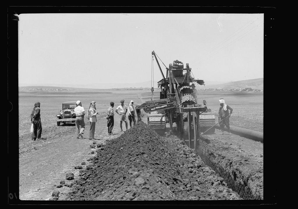 The image depicts a group of individuals engaged in agricultural activities, specifically the laying down of a pipeline. A large ditching machine is prominently featured at work on the barren ground, with several men around it observing or assisting. Some are using shovels to clear excess soil from the trench where the pipe has been laid.

A vintage automobile and an old tractor can be seen in the background, emphasizing the historical context of this scene. The landscape suggests a rural setting, possibly within Israel's plains near Nazareth foothills during July 1933 as part of Iraq Petroleum Company’s pipeline project across Esdraelon Plain.

The photograph is black-and-white and appears to have been taken by American Colony Photo Department with one negative on nitrate film measuring 4 x 5 inches.