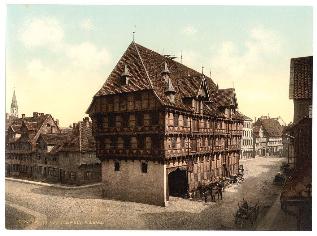 The image displays a historic European town scene, likely from the late 19th to early 20th century. The focal point is an imposing half-timbered building with several stories and steeply pitched roofs that are characteristic of medieval German architecture known as Fachwerkhäuser. This particular structure exhibits detailed timbering and a large arched entrance, indicative of its significance in the town's history.

The street in front shows horse-drawn carriages parked alongside or moving down it, suggesting this image captures daily life from an era before automobiles dominated transportation. Traditional houses with similar architectural styles flank one side of the main building, while on the other, there are modern buildings that imply a blending of old and new urban development.

In the background stands what appears to be a church spire or tower, signifying religious architecture's presence in the town layout. The sky is partially cloudy but mostly clear, hinting at fair weather conditions during this time period. There are no visible people, which adds to the stillness of the scene and emphasizes its historical nature.

The image includes text annotations indicating it was printed by P. Z. Braunshwigg., a company from Braunschweig (Braunschweig is another name for Brunswick), Germany between approximately 1890 and ca. 1900, suggesting that the photochrom print tec [...]