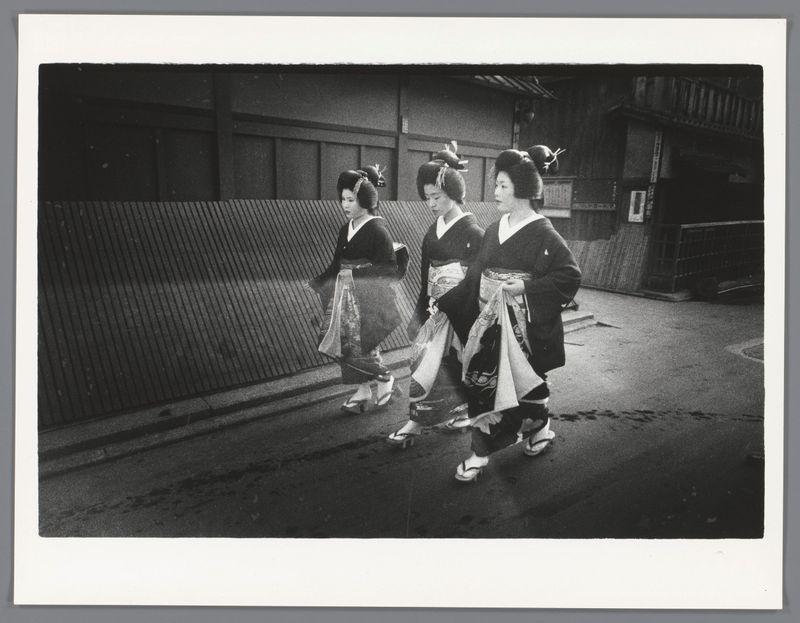 The image captures a moment on an urban street at night, featuring three individuals who appear to be geisha based on their traditional attire. The photo is in black and white, emphasizing the textures of clothing, skin, and surroundings with high contrast between light and shadow areas.

Geisha are traditionally dressed in elaborate kimonos called okāfan, which have wide sleeves known as fukuro-goi that extend past the waistline; this particular style suggests a kimono from the Edo period. Their hair is styled up into an intricate hairstyle adorned with decorative elements such as ornaments or flowers.

The geisha are walking side by side on what seems to be wet pavement, possibly after rain, which adds reflective qualities and enhances details in their garments and accessories like obi belts that tie at the waist. The choice of footwear includes traditional geta sandals made from wood for better grip on slippery surfaces.

Behind them is a backdrop featuring wooden structures with slatted partitions typical of urban Japanese architecture during mid-20th century periods, suggesting this might be an older district in Tokyo or another major city within Japan. There's no visible modernity except the streetlights casting shadows and illuminating parts of their path ahead.

Overall, it depicts cultural heritage amid contemporary life settings, perhaps hinting at a theme relat [...]