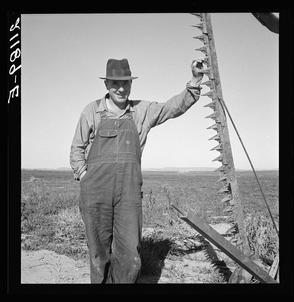 The image is a black and white photograph showing an individual outdoors in what appears to be rural or agricultural settings. The person, likely male based on clothing style and build, stands smiling at the camera with their right hand extended holding onto a metal object that has many pointed protrusions. They are wearing overalls, which suggests they may be engaged in manual labor or farming work.

The landscape behind them is open and dry, indicative of farmland or prairie-like terrain. There's no visible vegetation close to where the individual stands, but some sparse shrubbery can be seen further back. The sky above appears clear with no clouds, suggesting a sunny day which could affect visibility for color identification.

There are handwritten characters on the left-hand side that seem like they might indicate a date or an archive reference number, such as "7-2816." These markings suggest this photograph may be part of a larger collection. The person is wearing a hat and their clothing shows signs of wear which further supports the idea of them being engaged in labor-intensive work.

In terms of context provided by the caption linked to an image source (https://images.loener.nl/DorotheaLange/full/6495/6495b8afba25398d90c510a4.jpg), it indicates a connection with Mr. Browning and his activity in getting ready for mowing hay, situated possibly at Dead Ox Flat in Mal [...]
