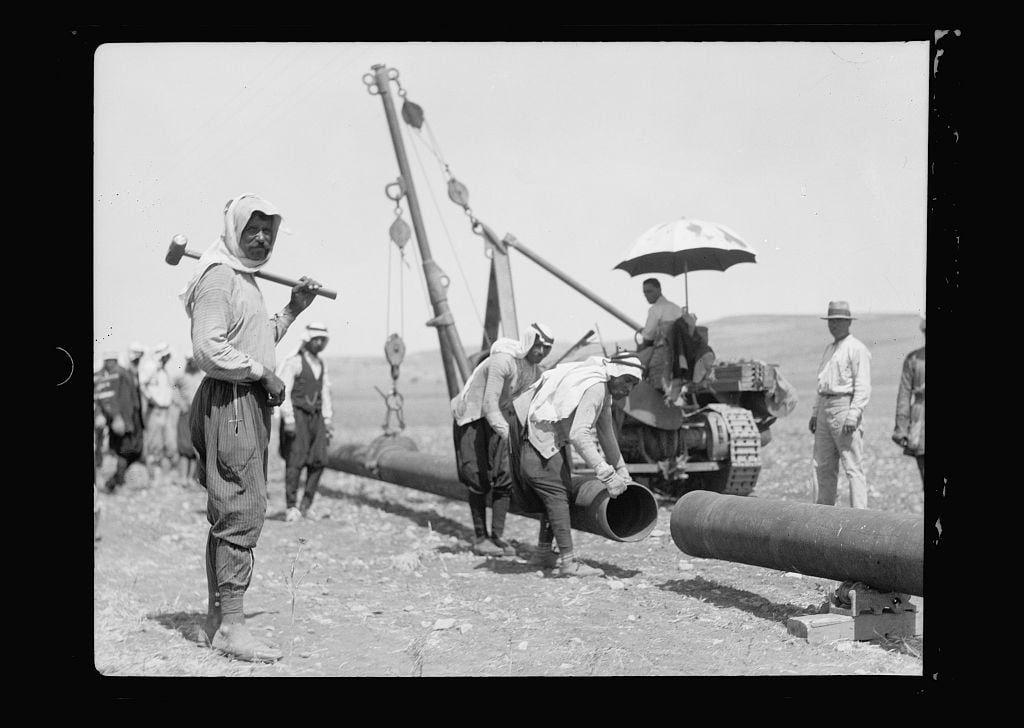 The image is a black and white photograph depicting a group of people working on the laying of a pipe line. The setting appears to be an arid landscape, likely in or around Jerusalem during July 1933 as part of Iraq Petroleum Company's pipeline project across Esdraelon Plain.
In the foreground, several individuals are actively engaged in assembling and installing large cylindrical pipes with tools such as hammers and shovels. One person is prominently featured holding a hammer over their shoulder while walking past the camera. Another individual appears to be using a shovel or brush-like tool on one of the pipes being laid out.
In the middle ground, more workers are visible around various pieces of heavy machinery including what seems to be a tractor crane used for lifting and positioning the pipe segments into place. The equipment is large in comparison to the people working with it, indicating its significant size relative to typical human strength or manual labor tools.
Amongst this scene stands an umbrella providing shade over one person who appears relaxed under the canopy of protection from direct sunlight exposure while observing others at work. This contrast suggests a mix of necessary labor and effort against environmental conditions like heat during that period in time, as well as offering comfort to those working hard outdoors on such hot days.
The background i [...]