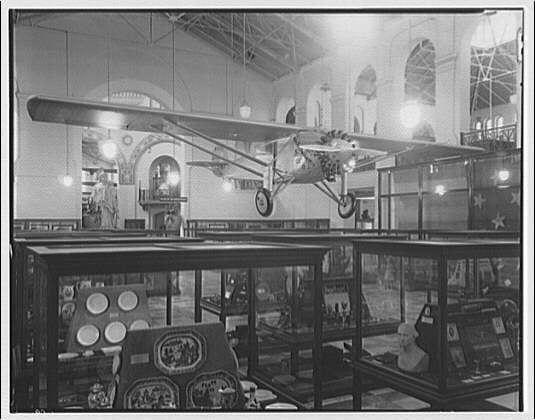 The image depicts an interior space, likely a museum or exhibition hall, with high ceilings and large arched windows. Prominently displayed is the Spirit of St. Louis aircraft, which appears to be suspended from the ceiling by wires. This airplane has two open cockpits and displays star insignia on its wings, indicative of military use.

Below the plane are various showcases containing artifacts such as plates with decorative designs, a bust statue resembling an animal or mythical creature, and other indistinct items that could possibly include historical memorabilia. The lighting is bright, suggesting overhead illumination typical for indoor exhibitions to highlight displays clearly.

The photograph appears black-and-white, which gives it a vintage feel consistent with the early 20th century timeframe suggested by the presence of the Spirit of St. Louis and its context within the Smithsonian Institution interiors reference provided in the caption.