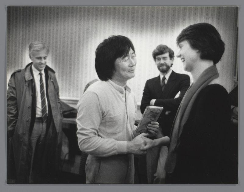 The image is a black and white photograph depicting four individuals in what appears to be an indoor setting. The central focus of the photo shows two women facing each other, engaged in conversation or exchange. One woman has short hair, wearing glasses, and dressed in a light-colored blouse; she holds some papers or documents with her hands clasped together as if presenting them.

To her right stands another smiling individual whose back is partially visible to the camera, suggesting they are standing close behind her. On the left side of the frame, there's an older man wearing glasses, and what looks like a trench coat over a suit; he has his arms crossed and appears contemplative or observant as if watching the interaction between the two women.

In the background stands another individual with short hair and a mustache who is also dressed in dark clothing. His posture indicates that he's standing slightly apart from the main group, possibly observing their exchange.

The setting looks like an office environment due to what appears to be blinds or curtains on windows behind them; however, no other specific details about this location are provided within the image itself. The overall mood of the photograph seems amicable and professional based on body language and facial expressions captured in it.