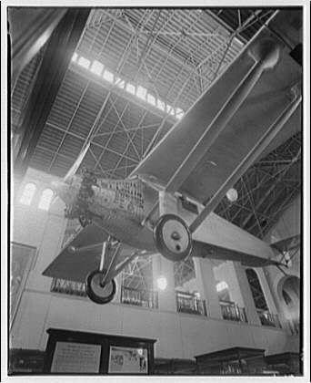 The image depicts an interior view of a large building with high ceilings and exposed beams, possibly part of a museum or exhibition hall. A vintage airplane is suspended from the ceiling, showcasing its undercarriage and propeller in detail. The photograph appears to be black and white, suggesting it could date back several decades ago. Surrounding areas include railings along balconies and informational signs on walls. This scene captures an artifact with historical significance, potentially connected to a notable aviation event or figure from the past century.
