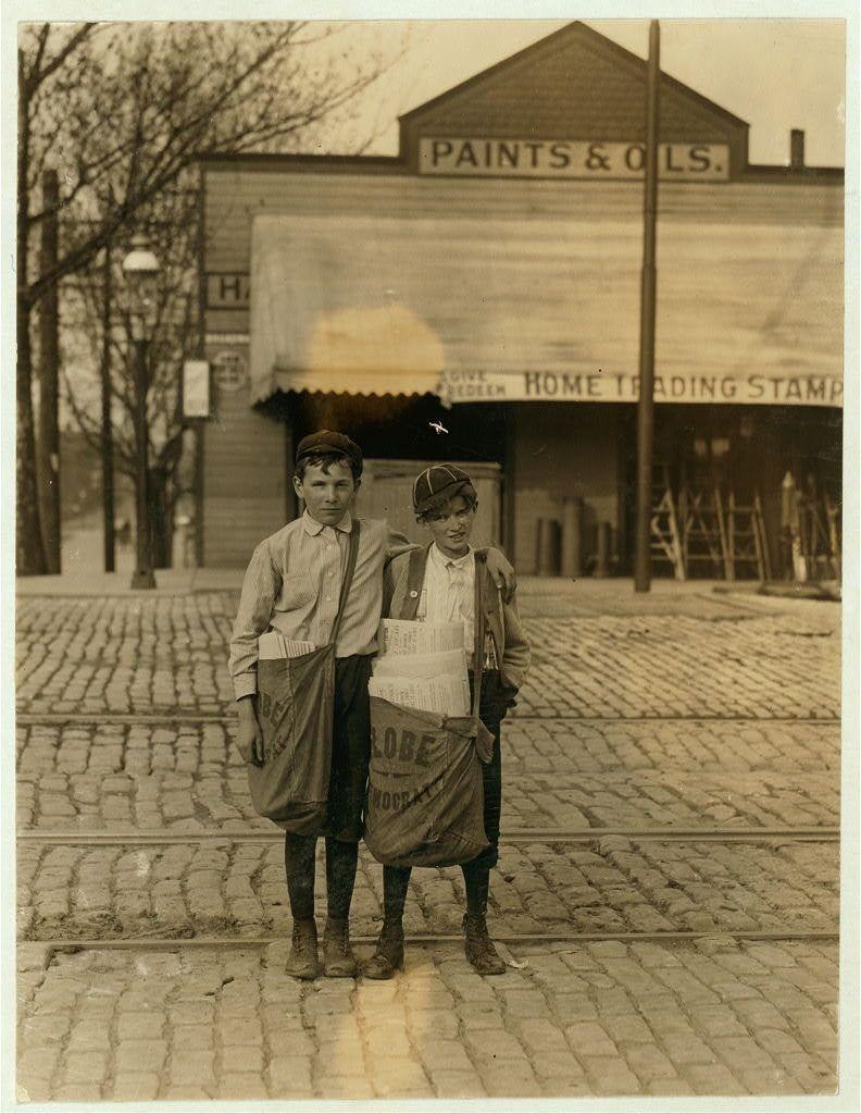 The image depicts two young boys standing on a cobblestone street in front of an establishment with the sign "PAINTS & OILS." The older boy is wearing striped pants, suspenders, and brown shoes, while the younger boy has on rolled-up trousers, dark boots, and what appears to be overalls. Both are dressed casually for their time period, which suggests early 20th century based on clothing styles. Their posture indicates they may have been posing specifically for this photograph as opposed to just being bystanders in a public space.

The establishment behind them features the words "HOME TRADING STAMP" suggesting it's possibly part of a cooperative organization or system where members receive stamps that can be exchanged for goods at participating stores, which was common during the early 1900s. The building has an industrial look with visible windows and ladders against its side.

The photograph is in sepia tone, indicating age as well as artistic choice by the photographer to create a vintage feel. There's no other people or moving objects captured here; it feels still and quiet. Trees without leaves suggest that this photo might have been taken during winter months which could correlate with historical events around those times.

The photograph is labeled "The boy with bag, nicknamed Turk, said he was going to Texas soon." The investigator found him recently with $1.75 he [...]