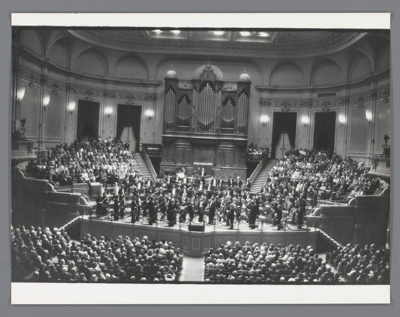 The image depicts a black and white photograph of an orchestra performing in a concert hall. The musicians, dressed in formal attire with many wearing dark suits and some in tuxedos or ceremonial dress, are seated on a large stage facing the audience which fills multiple levels of seating. They play various instruments such as violins, cellos, trumpets, and woodwinds among others.

In front of them is a grand organ with two large cases flanking either side, indicative of classical music venues like those found in Europe or North America during that era. The architecture suggests an older building possibly from the early to mid-20th century given its design elements such as arches and ornate detailing on the walls.

The audience is seated facing the stage with many individuals looking forward attentively, some turning their heads towards each other conversing casually in what appears a semi-casual environment despite being at an orchestral performance. The lighting seems to be coming from above, illuminating both performers and spectators alike creating soft shadows on the orchestra members.

The photo's composition captures not only the spectacle of music making but also reflects societal norms around public gatherings such as concerts during that period where formal attire was expected for all involved except perhaps some audience members who seem more relaxed in dress.