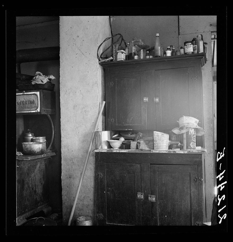 A black and white photograph depicts a cluttered kitchen with an old-fashioned stove, wooden cabinets filled with various bottles of condiments or medicine, bowls on the counter, and a collection of hats. A broom leans against one wall near some cans that appear to be empty paint containers. The setting suggests it may have been taken in an older home from an earlier era when such stoves were common, possibly reflecting rural conditions during the early 20th century or later periods depending on the exact location and context provided by additional information like a caption number as referenced here ("General caption number 72").