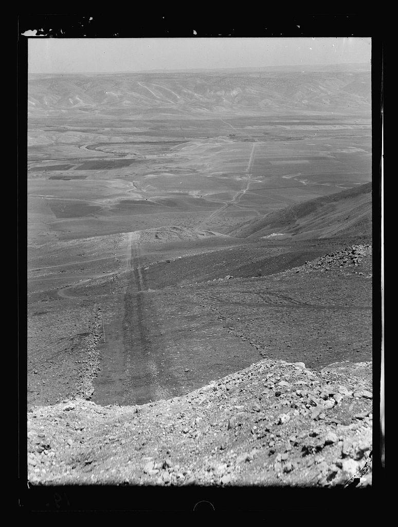 The image depicts a vast, barren landscape viewed from an elevated perspective. The terrain is rugged and uneven with prominent ridges and valleys that stretch out into the distance. A winding dirt path or road cuts across the middle of the scene, suggesting human activity in this remote area. In the foreground, there's a pile of rocks on what appears to be rocky ground. This black-and-white photograph has an aged quality, indicating it may have been taken several decades ago. The caption and additional context suggest that the image is related to the laying of Iraq Petroleum Company’s pipeline across the Plain of Esdraelon in July 1933, specifically showing a pipe line crossing through Jordan Valley from Beisan hills looking eastward. This photograph was likely part of a series documenting various aspects of early petroleum exploration and infrastructure development by this company during that period.
