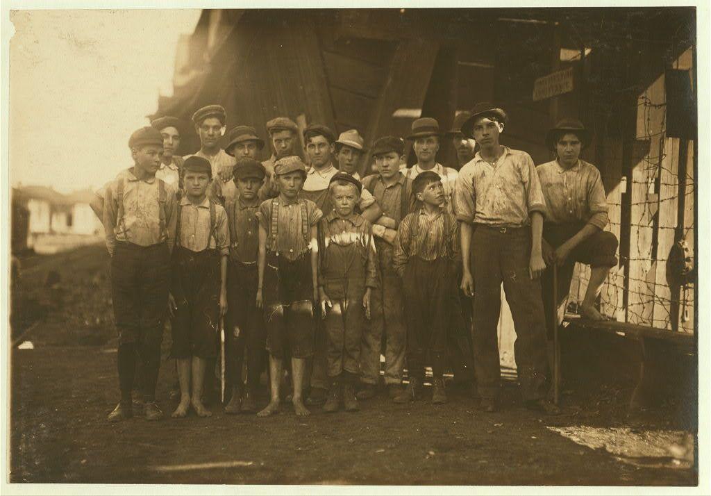 This black and white photograph depicts a group of young boys dressed in work attire from the early 1900s. They stand together on what appears to be a dirt path, with some wearing hats such as flat caps or newsboy caps and others without head coverings. Their clothing varies; many wear overalls, while several are clad in shirts tucked into high-waisted trousers. Some boys have suspenders visible under their outerwear. The group is posed for the photo, standing upright facing the camera with a range of expressions from serious to slight smiles.

Behind them, part of an industrial structure and barbed wire fence suggest they may be near a factory or mill setting. There's also partial signage that might indicate it’s related to 'Avondale Mills' in Birmingham, Alabama—a reference likely due to historical context provided by the image source mentioning "Avondale Mills" and referencing Lewis Hine.

The boys seem of varying ages but all appear young enough to be considered children or adolescents. It's unclear whether they are employed there voluntarily as laborers or if this photo represents a documentary aspect, perhaps capturing child workers in an industrial setting during that era. The historical significance lies not only in the representation of youth and their environments from around 1907 but also serves as evidence for early social reform movements against child labor  [...]