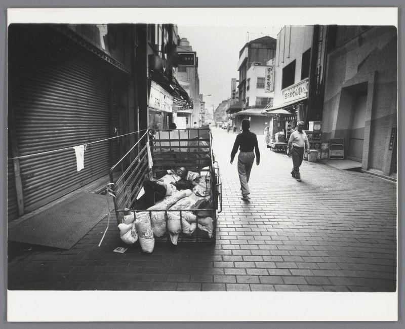 The image depicts a street scene in black and white, likely from an urban area. In the foreground on the left side of the photo is a large cart filled with what appears to be sacks or bags; some are covered partially by fabric that seems makeshift. The sack closest to us has been draped over another one like it, creating a sense of depth.

In the middle ground, two figures can be seen walking down the street: on the right side there's an individual wearing dark clothing and carrying a bag, seemingly in motion; this person is not clearly visible due to their stance. To the left, closer to the cart, another figure appears to lie inside it, covered with a blanket or piece of cloth that drapes over them.

The street itself is made up of brick paving stones and extends into the distance where other buildings loom on either side. Some have signage in what seems like Japanese script, indicating this may be in Japan as suggested by the caption provided for this image from an external source.

On the right-hand sidewalk there are various objects scattered: a small pile to one side appears discarded or left out and near it is some kind of machinery with a box on top. Further down, another figure walks away, appearing smaller due to distance but also wearing light-colored clothing that contrasts with their headgear—a cap.

The architecture suggests an older part of the city as moder [...]