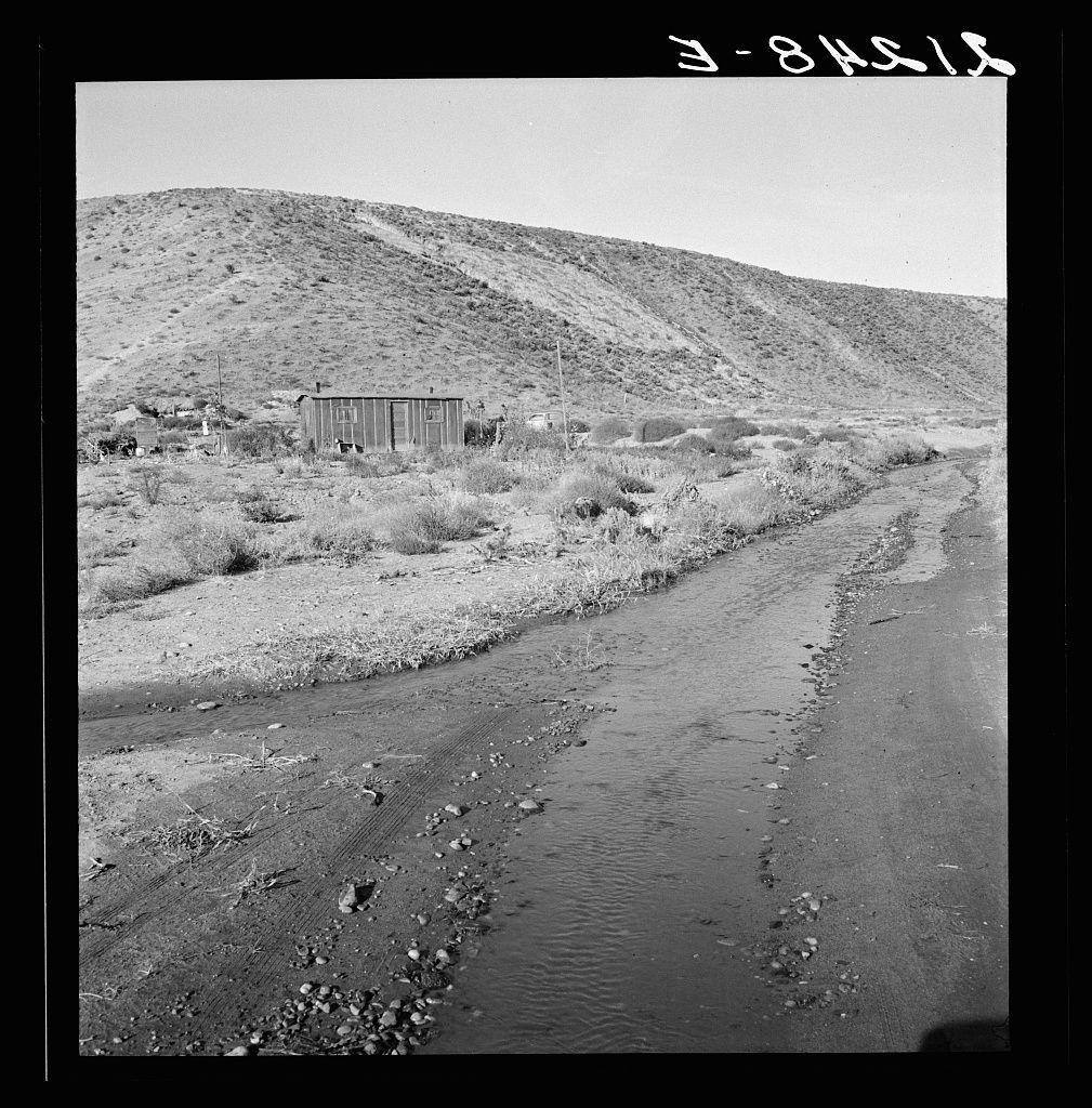 The image depicts a black and white photograph of an arid, rural landscape. A small wooden shack with boarded windows sits in the center foreground near a dry riverbed or creek bed that meanders through the scene. Sparse vegetation dots the area around the shack, indicating sparse ground cover typical of semi-arid regions.

A road winds up to the right from where the photograph is taken, leading away toward a hillside covered with rocky terrain and shrubbery under an open sky. The overall tone suggests desolation or hardship, which aligns with the caption mentioning that it's "awful slow" for those living in such conditions as described.

Additional context provided includes information about seepage of irrigation water affecting road conditions leading to a new farm, suggesting ongoing agricultural activity despite challenges faced by residents like the family who live in this shack. The specific location is identified as Dead Ox Flat, Malheur County, Oregon with reference number 67-1 indicating it might be part of an archived collection or series documenting rural life during that period.