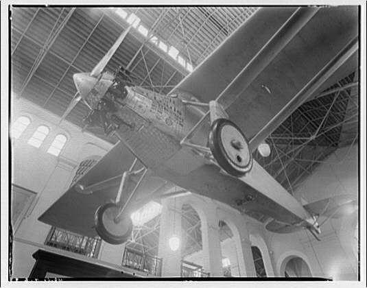 The image depicts a large, vintage biplane suspended from the ceiling of an indoor space with arched windows and ornate details. The plane is identified as "Spirit of St. Louis," which was famously piloted by Charles Lindbergh in his transatlantic flight across the Atlantic Ocean in 1927.

The photograph appears to be a black-and-white negative, likely taken around or after World War II (circa 1950), based on its style and quality. The image captures the plane's wingspan as it is displayed indoors, possibly within a museum setting given the architectural features in the background.