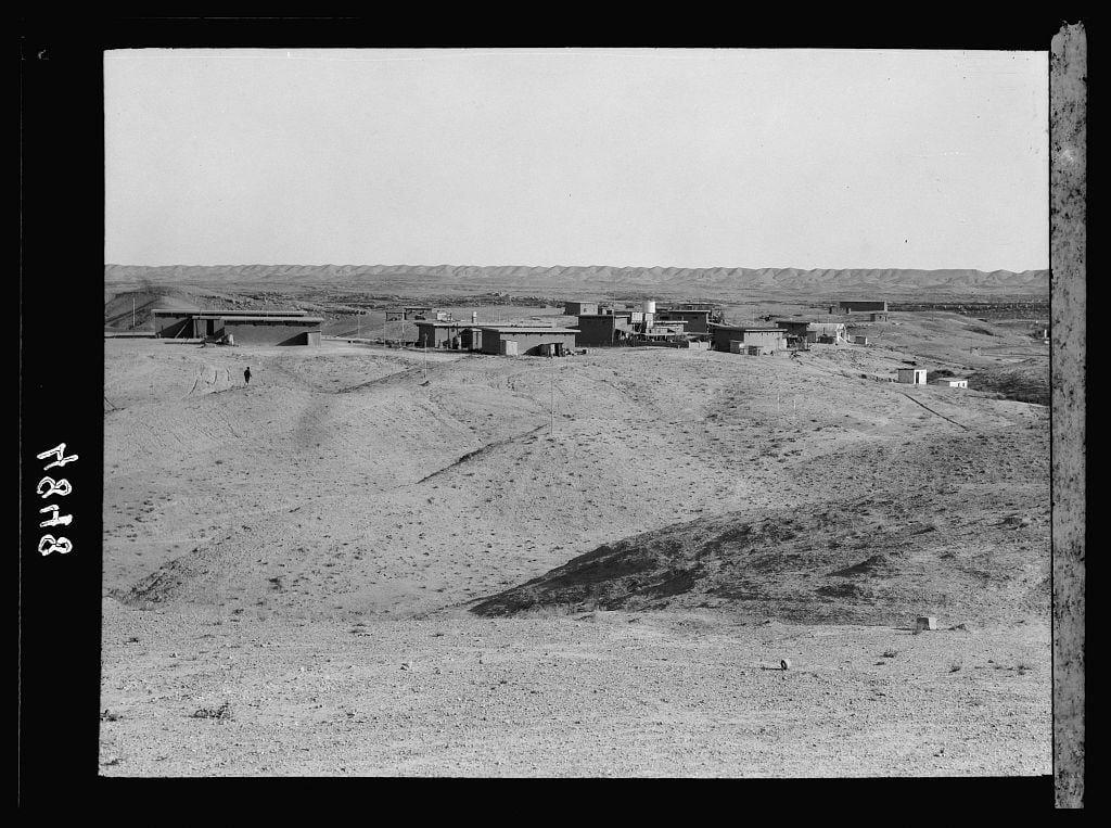 The image displays a black and white photograph of an expansive, arid landscape with minimal vegetation. In the middle distance, there is a cluster of industrial buildings that resemble oil extraction facilities or related operations. These structures appear to be made from concrete and have flat roofs, suggesting they might serve functional purposes rather than residential ones.

Scattered across the terrain are several smaller outbuildings and what could possibly be storage tanks or other equipment typically associated with petroleum production sites. The area looks barren, consisting mainly of sandy soil interspersed with sparse patches of grass and shrubs, indicative of a desert environment.

In the foreground on the left side, there is an individual walking away from the viewer's perspective towards these industrial buildings, providing a sense of scale to the vast landscape. On the right-hand edge, part of what seems like a wooden pole or beam can be seen, adding another element that suggests human activity and infrastructure in this remote area.

The terrain slopes gently upwards as it extends into the distance where faint lines suggest a mountain range on the horizon under a clear sky. The photo is labeled with "1843" which could possibly indicate either a location code or perhaps an internal reference within its collection, like that of an album number.

This sce [...]