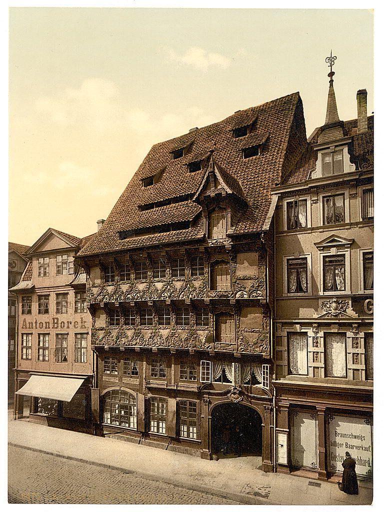 This image shows a historical European building, specifically in Brunswick (Braunschweig), Germany. The architecture is traditional with detailed ornamental woodwork and gabled roofs typical of the region's medieval buildings. A sign reads "Anton Borek," likely indicating either a business or residence name from that era. Another section bears signs for businesses like "Gebrauschkloge" which could suggest a maker of metal bells, brassware, and other items associated with bell making. The street is paved in cobblestones typical of older European cities, adding to the historic ambiance. A figure can be seen walking on the sidewalk at the bottom right corner of the image, dressed in period clothing that blends into the historical context depicted by the architecture around them.