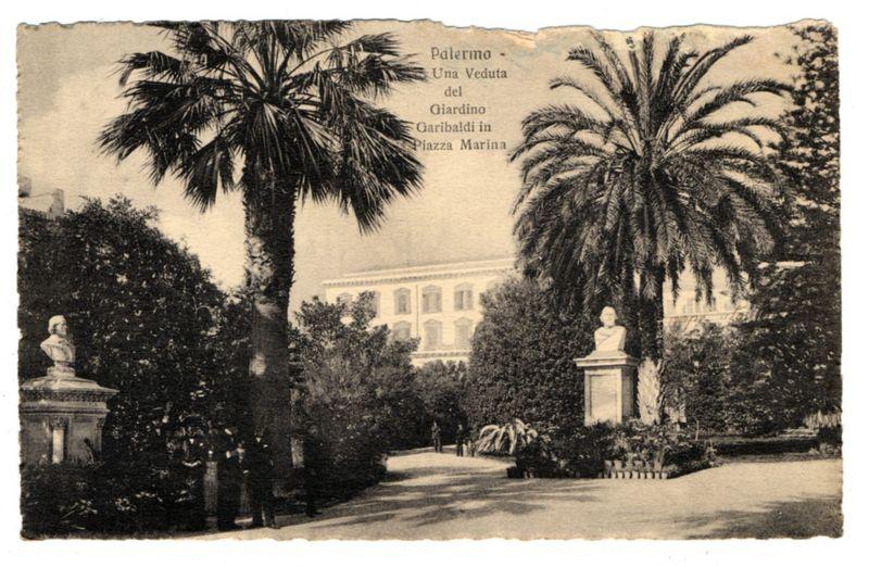 The image is a black and white vintage postcard depicting an outdoor garden setting. Prominent in the scene are tall palm trees framing the view of a classical-style building with visible windows and columns, indicative of neoclassical architecture. In front of this structure stands a statue on top of what appears to be a pedestal or monument. The foreground shows more greenery including bushes and shrubs, suggesting an ornate garden layout typical of historical estates.

There are two busts displayed prominently in the scene; one is placed slightly left in the frame while another larger figure towers over it further right. These sculptures add a sense of cultural significance to the setting. Two individuals can be seen walking along what appears to be a pathway, suggesting that this garden may also serve as a public park or promenade.

The text "Palermo - Una Veduta del Giardino Garibaldi in Piazza Marina" is printed across the top portion of the postcard, indicating that it's an image from Palermo showcasing views within Garden Garibaldi located at Marina Square. The paper shows signs of age and wear, including creases and discoloration around the edges, giving a sense of its historical value as a keepsake or for travel documentation.

Additional information about this specific postcard can be found on "https://images.loener.nl/FerroCandilera/full/6730/6730a60789b10e7a2 [...]