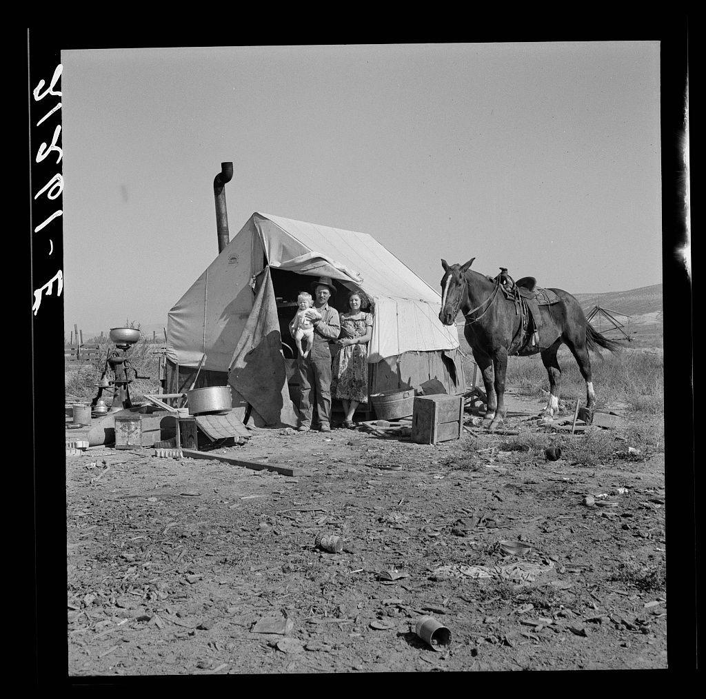 The image is a black and white photograph depicting a rural scene. In the center, there's a family gathered around a makeshift tent with one person standing inside holding an infant while another holds a young child outside by the entrance of the tent. Beside them to their right stands a horse attached to a wagon or cart that has not been fully visible in this angle. Various items are scattered on the ground near the campsite, including pots and pans likely used for cooking, barrels possibly containing water or other liquids, wooden crates which may be holding supplies like food or tools, as well as some loose debris from what appears to have once been a more organized setting that is now disarrayed. The terrain around them looks dry with sparse vegetation suggesting an arid climate typical of Oregon's high desert regions. In the background, there are faint outlines suggestive of rolling hills and other structures not clearly defined due to distance or focus on the main subject at hand.

The photograph seems like it could be part of a larger series documenting life during hard times in rural America; perhaps related to agricultural struggles or migrations common in the early 20th century. The presence of the horse, wagon, and makeshift tent suggests mobility often required by farm families moving from place to place following harvests or seasonal work demands.

This image [...]