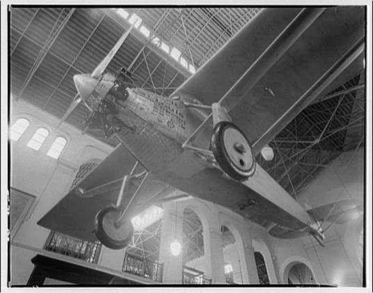 The image depicts a large, vintage airplane suspended from the ceiling of an indoor structure with arched windows and high ceilings. The aircraft appears to be a propeller-driven model with its landing gear extended on one side visible in the frame. It is displayed prominently within what looks like a museum or exhibition hall, given the architectural features such as arches and skylights that allow natural light to illuminate the space.

The airplane's body shows signs of wear and discoloration indicative of age, suggesting it may be an early model from around 1920. The propeller is in motion, implying either a subtle wind or perhaps some sort of mechanical activation within the display setup. There are no visible people or other objects that could indicate scale; however, the size relative to the architecture suggests it's quite large.

The black and white tone of the photo adds to its historical ambiance, indicating this photograph is likely from an earlier era when color photography was not as prevalent or widely accessible. The image captures a moment in aviation history, potentially showcasing one of the early aircraft that played significant roles during the early years of flight technology development.