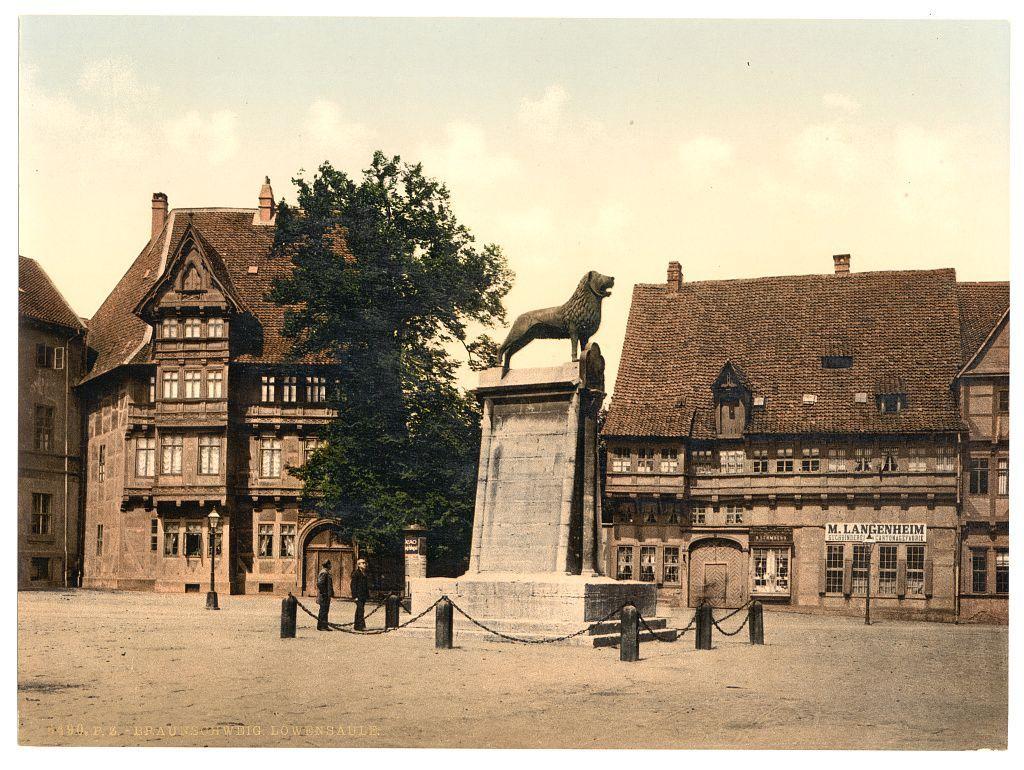 The image depicts a historical European town square, showcasing traditional architecture with timber-framed buildings. Dominating the scene is an imposing bronze statue of a dog on top of a pedestal, which appears to be in mid-leap or stride, suggesting motion and vitality. The sculpture stands as if leaping from one building's rooftop into another across what seems like a wide courtyard paved with large stones.

Around this central monument are various elements that contribute to the historical ambiance: aged lampposts lined up along pathways, metal fences demarcating certain areas or properties, and an open space where several individuals can be seen walking. The attire of these figures suggests early 20th-century fashion, reinforcing the historic context.

The buildings exhibit a range of details that highlight their antiquity; some feature prominent gables, dormer windows, decorative trimmings on wooden facades, as well as signage such as "M. LANGENHEIM" indicating local businesses or establishments from this era.

Inscriptions at the bottom left and right corners provide context: "BRABUSCHWINDLIG LOWENSÄULE," likely translating to a reference related to Brunswick's Lowenstraße (lion street), with an indication of 1890, suggesting that these are postcards. The overall composition captures not just architectural beauty but also offers a glimpse into the daily life and  [...]