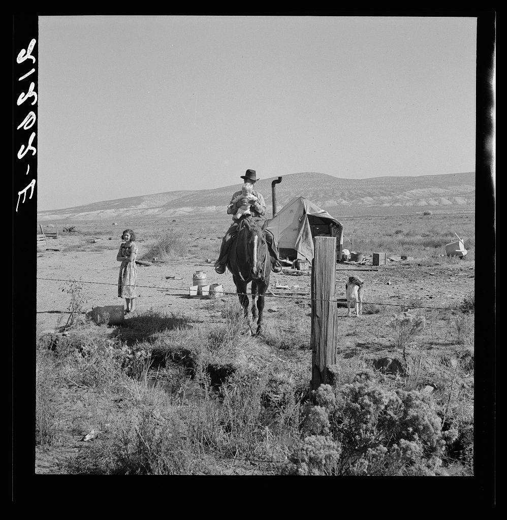 The image is a black and white photograph depicting an outdoor rural scene. In the foreground, there's a woman standing to the left wearing a knee-length dress with her arms crossed over her chest. She appears contemplative or waiting for something. To the right of the frame stands another young girl dressed in light-colored clothing holding onto what seems like a small object.

In the center and background is an older male figure riding a horse, possibly heading towards a makeshift campsite on his left side which includes various objects such as barrels, utensils, and buckets suggesting daily life activities. Behind him extends a desolate landscape with barren hills under a clear sky.

The setting suggests rural America during what might be the Great Depression era due to its simplicity and lack of modern amenities visible in this scene.
