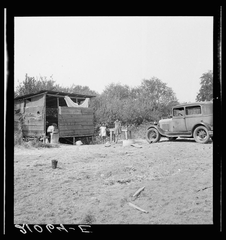 The image is a black and white photograph depicting an outdoor rural scene. There's a wooden shack with its side wall open, revealing a person inside who appears to be attending to something within the structure. A woman stands beside the door of the shack while another child sits on steps nearby. To the right, there's a vintage car parked next to what seems like farm equipment or possibly a large container. The ground is covered in dirt with some sparse grass and scattered debris such as sticks and metal objects. Trees are visible in the background under an overcast sky. There are handwritten markings at the bottom left corner of the photograph that read "DIOHN-E"