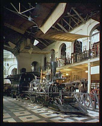 The image depicts a vintage railroad exhibit, likely located within the Smithsonian Institution Building around 1920 to 1950. This setting features an indoor space with arched windows and intricate structural details such as beams, ironwork, and wooden railings. The focus is on various locomotives and railway equipment displayed prominently in a large hall or museum area.
At the center of this exhibit stands a sizable steam locomotive with prominent smokestacks, wheels, and mechanical components visible underneath it, showcasing its intricate engineering design from an earlier era. Another vintage train can be seen partially obscured behind the foreground display piece. The flooring is patterned with white square tiles on top of what appears to be wooden planks.
The overall atmosphere conveys a sense of historical importance and industrial progress during that period in American history when railroads played a crucial role in transportation, commerce, and expansion across the nation.