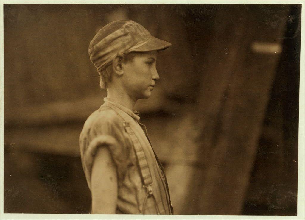 A sepia-toned photograph of a young boy standing in profile view against an indistinct background. The child appears to be wearing work clothes, including overalls and suspenders, with his right arm casually hanging down by his side. He is also sporting a cap that seems slightly askew on his head. His expression is solemn as he gazes off into the distance out of frame, suggesting contemplation or fatigue.

The photograph has an old-fashioned quality indicative of early 20th-century imagery and may be part of historical documentation related to child labor during that era in Birmingham, Alabama at Avondale Mills, a textile factory. The image is marked with visible damage such as scratches and discolorations throughout the print, which adds to its antique feel.

For more context on this specific photograph or similar works by Lewis Hine, one could refer to resources like https://images.loener.nl/LewisHine/full/6495/64955b4dba25398d90c339bb.jpg.