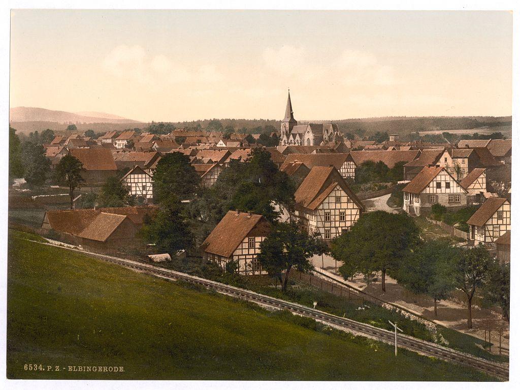 This image depicts a picturesque European village, likely from the late 19th century given its style and composition. The scene is dominated by traditional half-timbered houses with steeply pitched roofs that suggest they are built to withstand heavy snowfall or rain common in this region. These buildings have whitewashed walls between exposed wooden beams which was a popular architectural method, especially during the medieval period.

The village sits on gently rolling hills and appears well-integrated into its natural environment, surrounded by trees and open fields indicative of rural life at that time. A church with a tall spire punctuates the skyline in the background, suggesting it's an important local landmark or center for community gatherings.

A railway track cuts through part of the scene, hinting at industrialization or modern transportation advances during this period. The train tracks are bordered by simple fencing and grassy embankments typical of early railways before modern construction techniques were prevalent.

The overall color tone is warm with sepia overtones suggesting age or that it's a print from an old photograph album. This image exudes a sense of peaceful rural life, possibly capturing the essence of Germanic village living during this era.