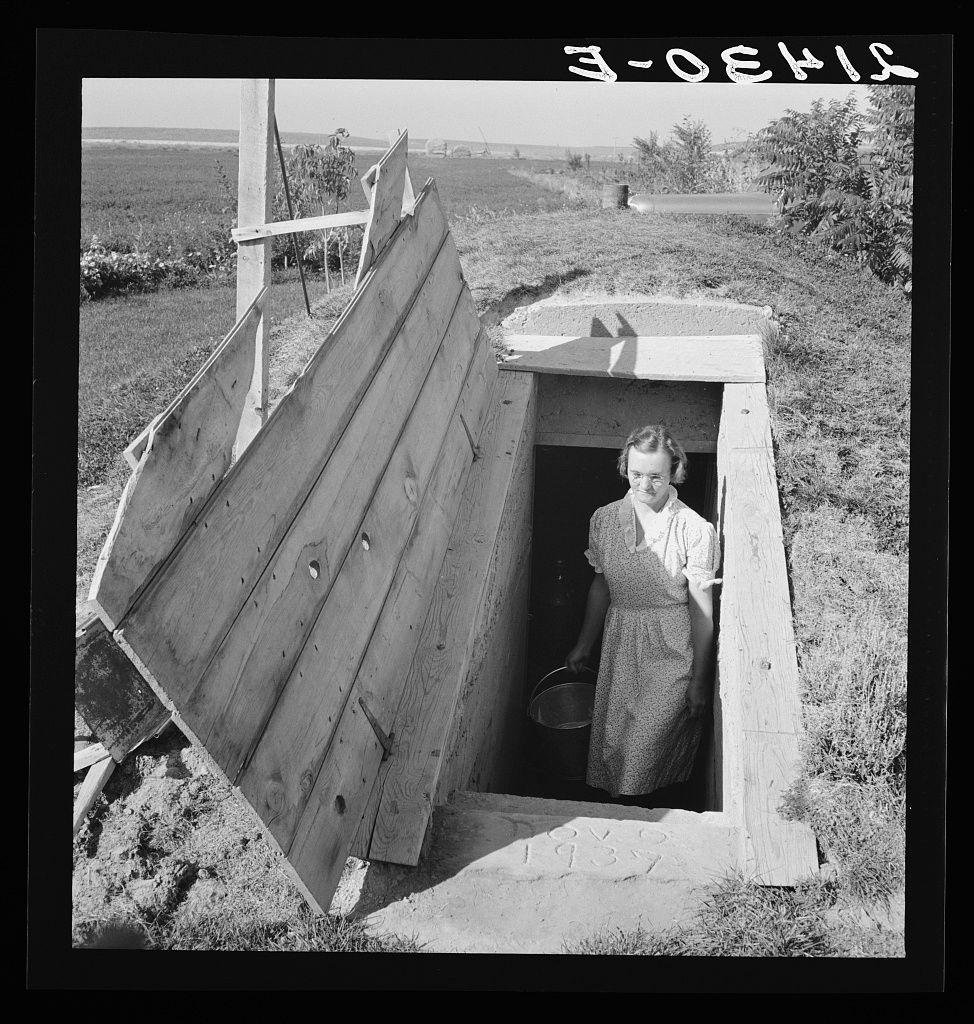 The image captures a moment in what appears to be rural life from the early 20th century, as evidenced by the black and white format. A person is emerging from an open wooden structure identified as a storage cellar on the Botner farm located near Nyssa Heights in Malheur County, Oregon. The individual wears a long-sleeved dress with puffed sleeves and what seems to be suspenders or decorative elements across their chest, paired with dark shoes that suggest practical footwear for working outdoors. They are holding a metal bucket in one hand, suggesting they may have been retrieving something from the cellar or preparing to do so.

The setting is rural and open, indicating an agricultural environment typical of mid-century Oregon farming communities. In the background, we can see crops extending into the distance under a clear sky with minimal cloud cover. A few trees are visible in the far right corner, while closer by, flowering plants add color contrast against the otherwise monochrome photograph.

The cellar itself is constructed from large wooden planks and features an open hatch on top that has been propped up to allow access for its use as a storage space. The ground leading into the cellar appears to be compacted dirt or gravel, with no signs of modern pavement work. A metal ladder leaning against the structure provides additional context about how one might climb  [...]