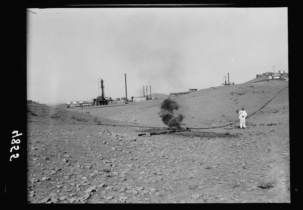 The black and white photograph depicts a rugged, rocky landscape with an industrial facility in the background. The foreground shows sparse vegetation on the barren hillside. In the center of the image is a large cloud of smoke or steam rising into the sky from what appears to be an oil well or gas flare. A single person dressed in light-colored clothing stands near this source of emission, holding onto some sort of equipment that may be part of the operation controlling or extinguishing the flame.

The scene suggests an early 20th-century industrial setting, possibly related to the Iraq Petroleum Company (IPC) mentioned in a caption. The IPC was involved with oil extraction and refining operations during its active years from around 1927 until it ceased operating there due to political instability following World War II.

Additional context for this photograph may be found within resources about American Colony's photo department, which could have documented various aspects of life or industry in the region. The image is an early negative on nitrate film and measures four inches by five inches in size (4 x 5 inch).