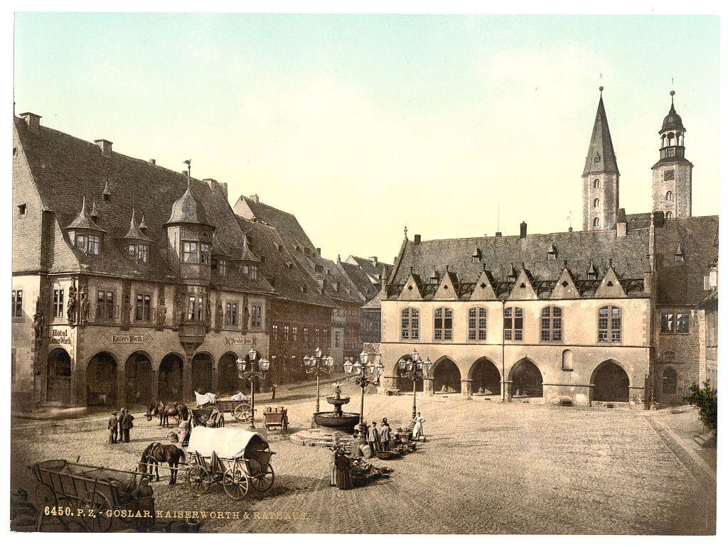 The image depicts a historical European town square, likely from the late 19th or early 20th century. It shows an open area with cobblestone paving and several horse-drawn carriages scattered around. In the foreground, there is a large carriage pulled by two horses, surrounded by people engaged in various activities such as conversing and tending to goods on display.

On one side of the square stands a building labeled "Hotel P.Z." indicating it might be a hotel or inn associated with P.C. Goslar. Another sign reads "Keller's War," which could refer to an underground cellar for wine storage, suggesting that this establishment may have been used as a tavern or restaurant.

The background features more buildings with traditional European architecture, including steeply pitched roofs and dormer windows on the upper floors. A prominent building in the center has multiple arched openings, possibly serving various commercial purposes like shops or market stalls.

Two towers rise above the rooftops to the right side of the image, indicating a town hall or another significant municipal structure. The overall atmosphere is bustling yet orderly, reflecting daily life and commerce within this historical setting.