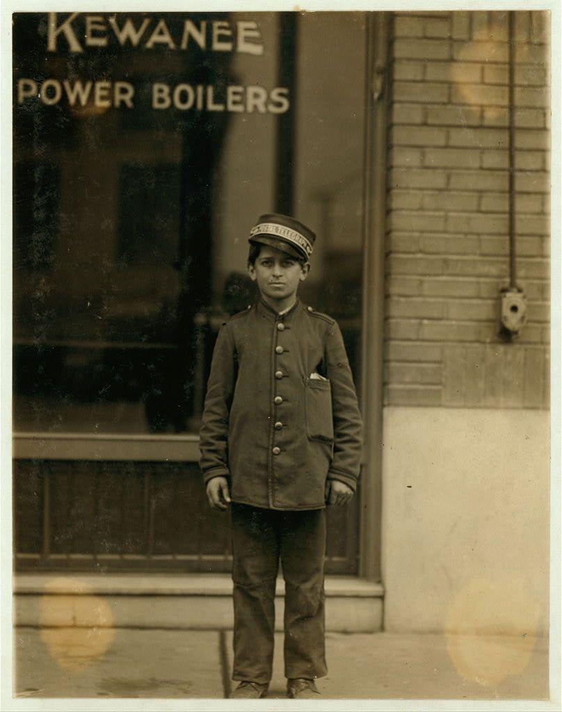 The image depicts a young boy standing in front of a building with signage that reads "Kewanee Power Boilers". The photograph is monochromatic and appears to be from an earlier time period, judging by the clothing style and overall aesthetic. The boy wears a uniform consisting of a dark-colored jacket adorned with multiple buttons down the front, two rows on either side, as well as epaulets on his shoulders, indicative of some formality or rank in service. He also sports a peaked cap that is characteristic of early 20th-century workwear for boys who were employed by companies such as railroads and utilities.

The boy's expression seems neutral to slightly somber, with direct eye contact towards the camera lens, suggesting that he may be familiar with having his photo taken or at least not opposed to it. His posture is upright but relaxed, with one hand partially inserted into a pocket of his jacket. The background suggests an urban environment due to the brick wall and what appears to be part of a utility box affixed on the exterior.

The building behind him has large glass windows through which additional details are obscured; however, "POWER BOILERS" is prominently displayed in capitalized letters at the top section visible above his head. The overall setting suggests that this photo was taken during work hours or possibly after such as part of a series documenting youn [...]