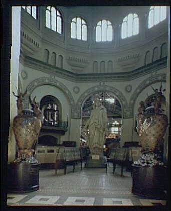 The image depicts a grand interior space, likely part of the Smithsonian Institution building. The focal point is a statue standing on an elevated platform surrounded by ornate urns with what appear to be deer heads as handles or decorative elements. There are several benches and display cases around the area for visitors to sit and view exhibits.

Several arched windows allow natural light into the space, which contributes to its airy atmosphere. The architecture includes classical details such as columns and intricate patterns on the walls and floors. Overall, it conveys a sense of historical significance and cultural importance associated with one of America's leading museums.