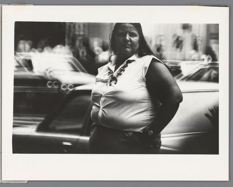 The image depicts a woman standing in an urban setting, with cars and city life blurred out behind her. She is dressed casually yet stylishly; wearing a sleeveless blouse adorned with decorative embellishments around the neckline which suggests cultural significance or personal style choice. The photo captures her mid-torso to full-length, indicating that she may be of average stature for an adult woman but it's not possible to determine precise measurements from this angle.

The monochromatic nature and quality suggest a vintage feel, with high contrast between light and shadow creating depth in the image. The focus is on the subject who appears confident as her hands are placed comfortably by her side. This photograph could be part of an exhibition or collection that documents urban life during its time period, potentially reflecting social dynamics and fashion trends from when it was taken.

Without additional context provided within this frame alone, more specific details such as age, race/ethnicity, clothing brand, exact location or event are not determinable. The watermark indicates the image may be held by an archive associated with Ed van der Elsken's work in Japan between 1959 and 1988 but beyond that date range no explicit copyright claim is visible within this snapshot alone.

It’s important to note ethical considerations when describing images of people, parti [...]