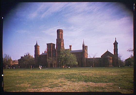 The image depicts the exterior of a grand historical building with multiple towers and spires, showcasing classical architectural features such as arched windows, decorative stone work, and tall chimneys. The structure appears to be made primarily of dark red brick or similar material, giving it a robust and imposing appearance.

The scene is set in an expansive grassy area that stretches out toward the building, suggesting open space possibly used for lawn activities or gatherings. There are some individuals visible at various distances from the camera, providing a sense of scale to the massive structure behind them.

A clear sky with few clouds occupies the upper part of the image, indicating fair weather conditions during the time when this photograph was taken. The colors in the photo have faded slightly over time due to exposure and age, giving it a vintage feel consistent with images from around 1920-1950 as mentioned.

In terms of context, the Smithsonian Institution is an American national research museum established by Congress on August 17, 1846. The building featured in this image could be part of its complex or related facilities, reflecting the architectural style often associated with government and educational institutions from that era.