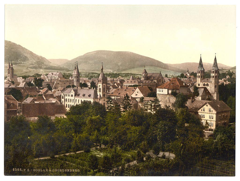 The image depicts a picturesque, historical European town nestled among rolling hills and lush greenery. The architecture is characterized by steeply pitched roofs, pointed spires of churches or towers reaching towards the sky, and an array of buildings with varying roof styles including tiled reds, shingles, and slate. Some structures exhibit half-timbering on their facades.

The town appears dense but organized, suggesting a well-planned urban layout typical for historic cities in Europe during its heyday. The coloration has the sepia-toned hue indicative of late 19th-century photographic techniques like photochroms or tintypes which were popular at that time.

In terms of flora, there are abundant trees and shrubs that create a dense green barrier between the town's edges and what seems to be cultivated garden spaces. These areas with structured hedges and rows suggest agricultural activity alongside urban development.

The photograph is labeled "6508.P.Z - GOSLAR&GEORGENDERG," indicating it may have been published or created by a photographer associated with Goslar, Germany; however, the specific dates provided range from 1890 to 1900. The landscape and architectural style could suggest this image is depicting Goslar itself as well as Georgenberg, possibly showing both areas in one view.

The overall feel of the photograph evokes a sense of nostalgia for an era when s [...]