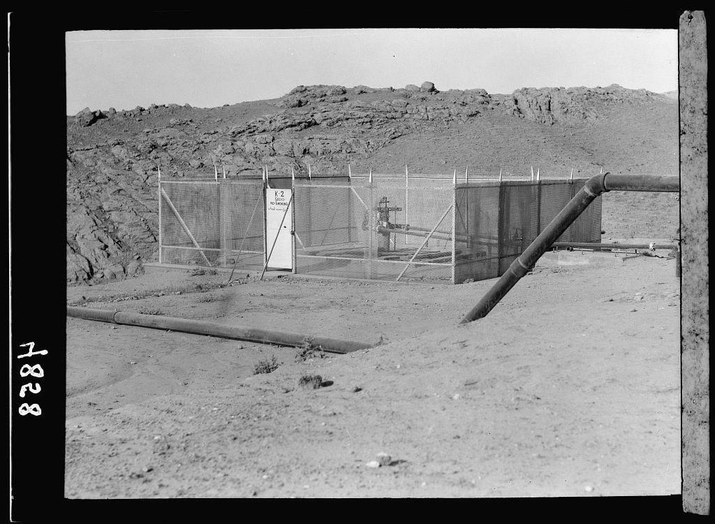The image is a black and white photograph depicting an oil well site. It shows dry, sandy terrain with scattered rocks and hills in the background under a clear sky. In the foreground, there's a large metal pipe lying on the ground running horizontally across the frame.

In the middle of the scene stands a fenced area containing various equipment such as storage tanks or filters for processing crude oil. A sign attached to one side of this enclosure reads "K-2," with additional text which is not clearly visible due to distance and resolution constraints. There are several pipes connected to the tank structure, suggesting an active extraction site.

The photograph appears dated, likely from a historical archive related to petroleum operations in Iraq during the early 1930s as indicated by its citation mentioning Kirkuk District and American Colony (Jerusalem) Photo Department. The image's metadata reference suggests it was taken on nitrate film with dimensions of approximately four inches wide by five inches high.

This photograph provides a glimpse into oil extraction activities from an earlier era, showcasing the infrastructure involved in such operations amidst arid landscapes typical to Middle Eastern regions where petroleum is prevalent.