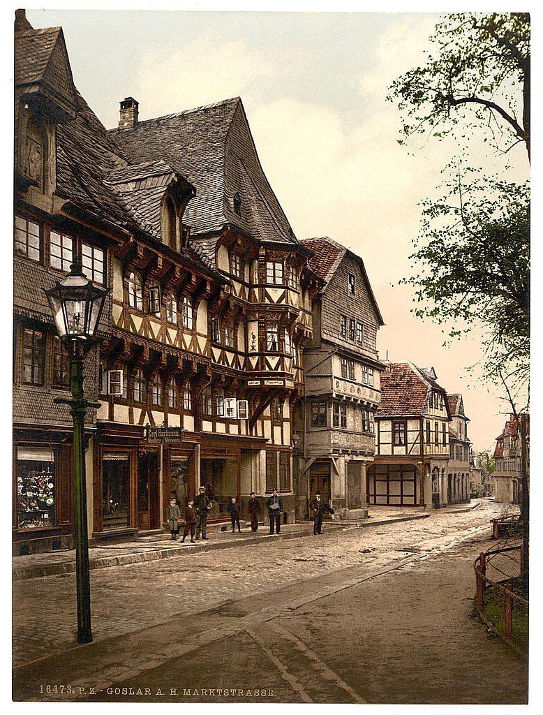 The image depicts a historic European street scene, likely from the late 19th or early 20th century. Traditional timber-framed buildings with steep gabled roofs line one side of the cobblestone road, which has horse-drawn carriages and pedestrians visible on it. The architecture is characterized by exposed wooden beams supporting the floors above ground level, often filled in with plasterwork to create a decorative effect known as "stuccowork." These structures have large windows with iron crossbars and feature various levels of balconies or galleries.

The street appears relatively narrow but stretches into the distance, flanked on both sides by buildings. Street lamps are visible along the curb side, indicating this was an urban area during that era. The sky is overcast, casting a soft light across the scene without harsh shadows. There's greenery with trees lining part of one building and what appears to be market stalls or shops at street level.

Signage on buildings suggests businesses such as cafes (possibly "Café Esmeraldau") which might offer refreshments like coffee. The overall atmosphere is serene, indicative of a small-town German setting in the Hartz region around Goslar during that time period.