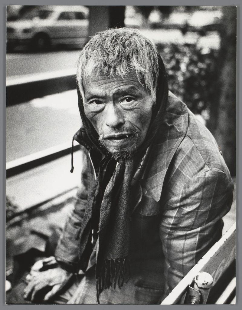 The image is a black and white photograph depicting an elderly man with weathered features, sitting on what appears to be public transportation. His attire consists of a plaid jacket over ragged clothes, suggesting he may be homeless or in need of resources. The texture and condition of his clothing indicate weariness and exposure to the elements.

His hair is disheveled, and he has visible wrinkles around his eyes, mouth, and forehead—characteristics that convey hardship and age. His hands are resting on a strap beside him; one hand appears slightly deformed or scarred. The man's expression could be interpreted as contemplative or weary. 

In the blurred background, we can discern an urban setting with vehicles passing by, which suggests movement and perhaps a busy street life beyond his immediate surroundings.

The photograph carries an air of documentary photography, capturing candid moments in everyday life without overt staging. It evokes themes of social awareness, human resilience, and possibly societal neglect or poverty amidst the hustle and bustle of city living.