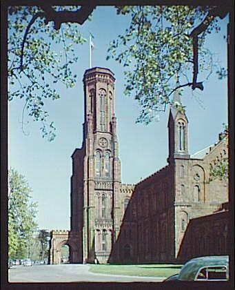 This image showcases the historic exterior of a grand, red-brick building with prominent architectural features such as arched windows and ornate detailing. The structure appears to be an institutional or governmental edifice, possibly dating from between 1920 and 1950 based on its style. A flagpole stands prominently at the topmost part of the tower, suggesting a place of significance. Surrounding greenery indicates that it is situated in an open area with trees nearby. The presence of vintage automobiles parked outside provides context for the time period when this photograph was likely taken.