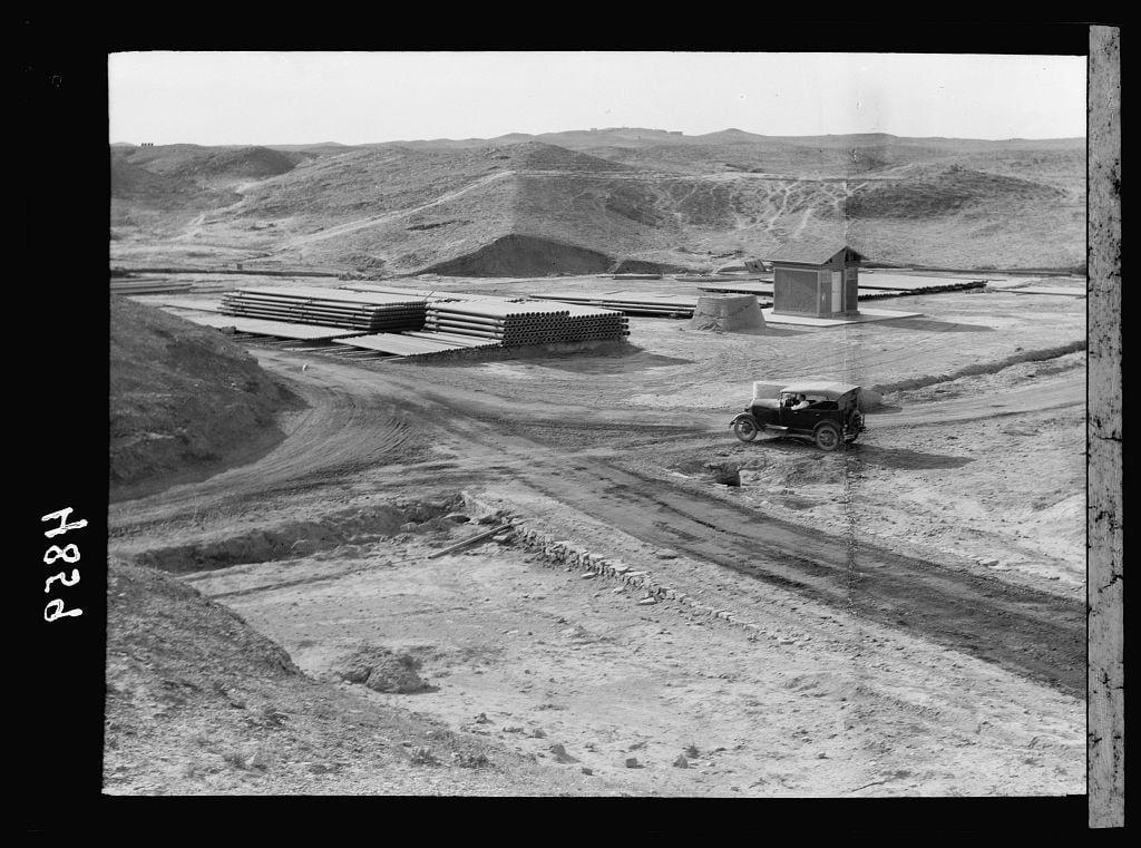 The image is a black and white photograph depicting an oil field site with various elements indicative of early 20th-century industrial activity. The landscape features undulating hills in the background, under a clear sky suggesting arid conditions.

In the foreground, there's a dirt road leading to scattered puddles reflecting some sunlight, likely from recent rain or mist. Alongside this path lies what appears to be constructed earthworks and broken stones, indicative of excavation work possibly related to oil extraction activities.

The middle ground is dominated by stacks of long cylindrical metal pipes aligned in neat rows extending into the distance. These are probably drilling equipment intended for use with oil wells. Adjacent to these, on a raised platform or embankment made from stone masonry and dirt layers, sits what looks like an open-air structure resembling a shed or storeroom.

A notable feature is a vintage automobile positioned close to one of the pipe stacks; it's likely used by workers for transportation within this site. The vehicle displays minimalistic design features typical of early automotive models with exposed wheels, simple headlamps, and rudimentary bodywork.

There are smaller structures in the vicinity including what appears to be storage units or possibly housing for personnel involved at this work site. In summary, it portrays a snapshot [...]