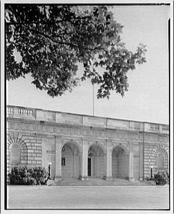 The image displays a black and white photograph of the exterior facade of what appears to be a neoclassical style building. The structure has multiple arches with columns supporting them, creating a colonnade along its length.

At the top left corner, there's an inscription or label that reads "B.D.P." which could possibly denote the photographer’s initials or some other identifier related to this image.
The setting is outdoors and it looks like the photograph was taken during daylight. There are trees with sprawling branches in front of the building adding a natural element to the otherwise architectural scene.

In terms of context, as indicated by additional information provided: This picture likely dates from around 1920-50 based on its appearance and given that Horydczak, presumably Theodor Horydczak according to other mentions in this dataset was active during approximately late 1800s - early mid-twentieth century. His works often feature detailed architectural subjects.

This photograph is part of a collection at the Freer Gallery which suggests it may have been taken as an official record or documentation, perhaps for artistic study purposes given Horydczak's background in photography and his work with architecture-related imagery.