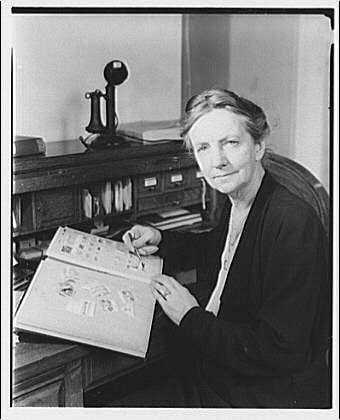 A black and white photograph depicting a woman seated at her desk, examining an open book with illustrations. The setting appears to be the Smithsonian Institution's National Museum of Natural History during its early years as a research museum. She is writing in the margins or on one of the pages while holding another page outstretched before her for reference.

The image captures Miss Manning from approximately 1920 to ca. 1950, and it was created by Theodor Horydczak (likely between around his birthdate - c.