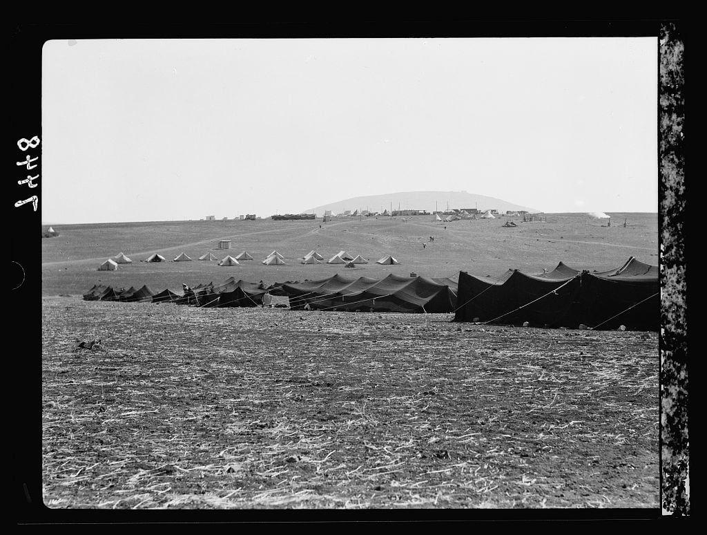 The image depicts a scene of numerous military tents spread across an open field. These large, dark-colored tent structures are arranged in rows and columns with some sparsely placed at the ends or along pathways between them. The terrain appears dry and barren with no vegetation visible on it.

In the distance behind these tents is a hilly landscape that gradually rises to form hills or small mountains. Atop one of these elevated areas, there seems to be an installation or settlement consisting of smaller structures, which could possibly indicate human habitation or some sort of military outpost. The sky above is clear and devoid of clouds.

This black-and-white photograph gives a historical feel, suggesting it might have been taken during wartime based on the arrangement of tents resembling that used by army encampments for soldiers' quarters, storage facilities, medical units etc.