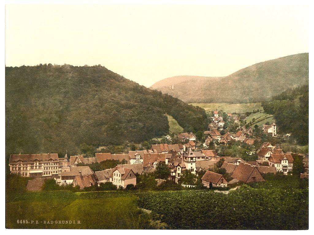 The image depicts a picturesque, rural village nestled amidst lush greenery and rolling hills. In the foreground, there's an expanse of verdant fields leading up to clusters of traditional European-style buildings with steeply pitched roofs. The architecture showcases timber-framed structures typical of certain regions in Europe, predominantly seen in Central or Northern parts of Germany.

The middle ground is dominated by a dense collection of houses and cottages that form the heart of this village landscape. Among them stands out larger, more elaborate constructions which could serve as community buildings like churches or town halls. The colors used for these structures are warm earth tones—reds, browns, and yellows—that blend harmoniously with their surroundings.

In the background lies a series of gentle hills densely covered in trees and shrubbery that fade into varying shades of green under an overcast sky, suggesting either early morning or late afternoon light. The overall atmosphere is peaceful, reflective perhaps of daily life around this time period as indicated by architectural styles which were prevalent between ca. 1890 to ca. 1900.

The caption indicates that the photograph was taken in Bad Grundl H., presumably a village located within Bath Grund (i.e. Bad Grund) region in Germany's Harz Mountains, during this historical period. The image is also identifi [...]